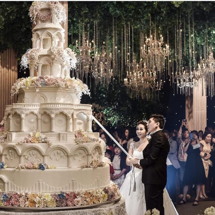 Novios sujetando una espada para cortar su pastel de bodas de 8 pisos decorado con betún blanco y flores de azúcar de diferentes colores