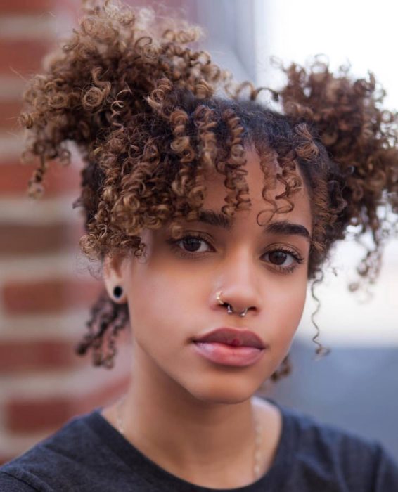 Ideas de peinados para el calor; mujer morena de cabello chino y castaño. con dos chongos altos y fleco, con piercing en la nariz y en el tabique