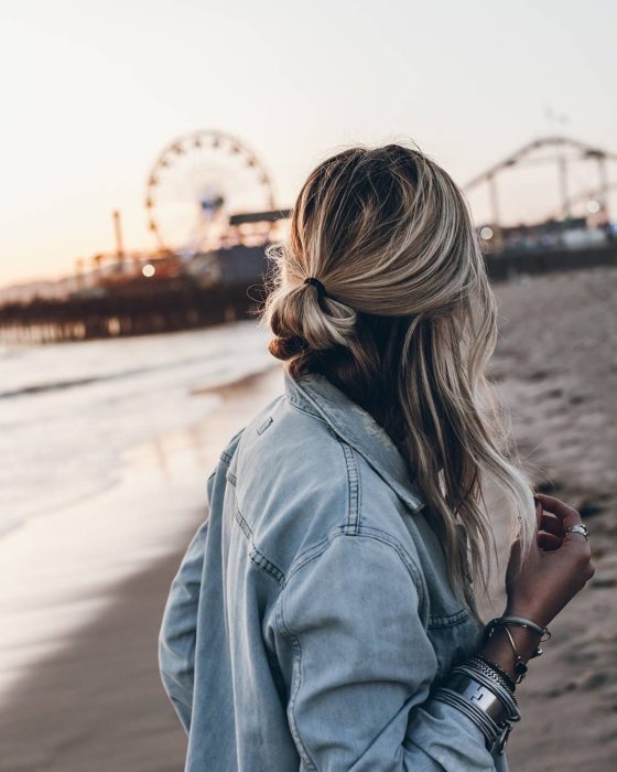 Ideas de peinados para el calor; mujer con cabello rubio ombré, peinada con media cola; en la playa con una rueda de la fortuna en el fondo