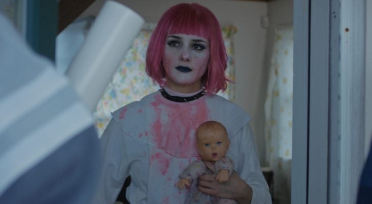 Película Little sister; chica con estilo gótico, cabello de honguito, con fleco, color rosa, usando maquillaje blanco y labial negro, sostiene en sus manos un muñeco