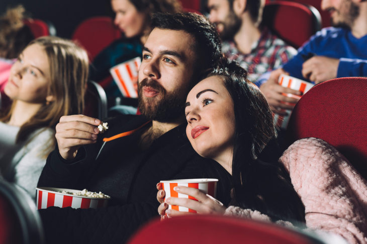 Chica recargada sobre el hombro de un chico, dentro de un cine comiendo palomitas