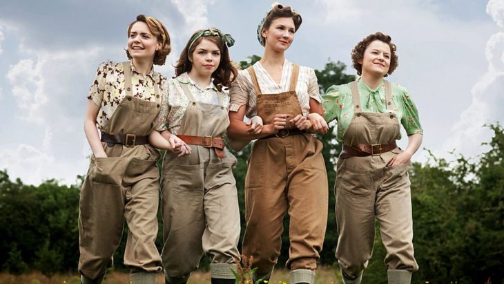 Escena de la serie de Netflix: The Land Girls. Mujeres vistiendo con overoles mientras caminan abrazadas 