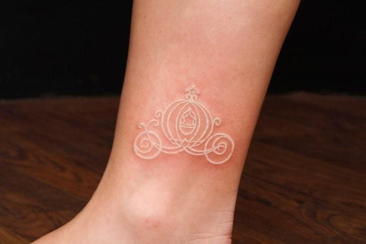 Tatuaje minimalista con tinta blanca del carruaje de Cenicienta en el tobillo