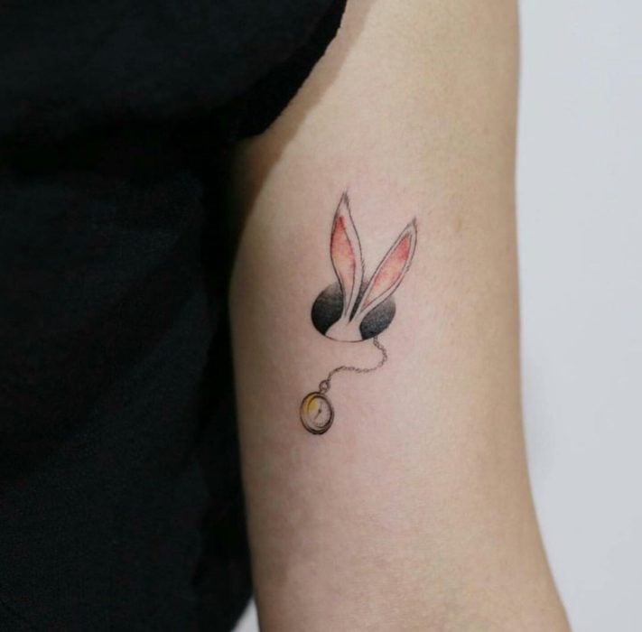 Tatuaje minimalista de las orejas y el reloj del conejo de Alicia en el País de las Maravillas en el brazo