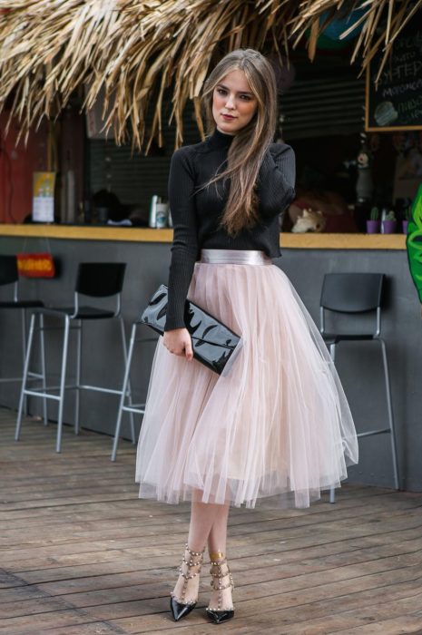 Ideas de vestidos para boda en jardín al aire libre; chica de cabello largo y suelto, con blusa negra, de cuello alto y mangas, con falda rosa de tul, bolsa negra de mano y tacones con estoperoles