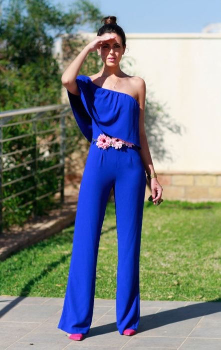 Ideas de vestidos para boda en jardín al aire libre; mujer de cabello castaño con peinado de chongo alto, tapándose el sol con la mano, usando jumpsuit azul rey largo, con cinturón de flores
