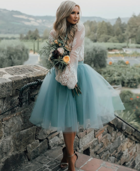 Ideas de vestidos para boda en jardín al aire libre; chica de cabello rubio platinado, largo y ondulado, con falsa de tul estilo bailarina de color azul, y top de encaje blanco, posando con un ramo de flores