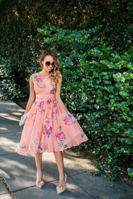 Ideas de vestidos para boda en jardín al aire libre; chica junto a una enredadera con vestido de tul rosa con flores, stilettos con moños y lentes de sol redondos