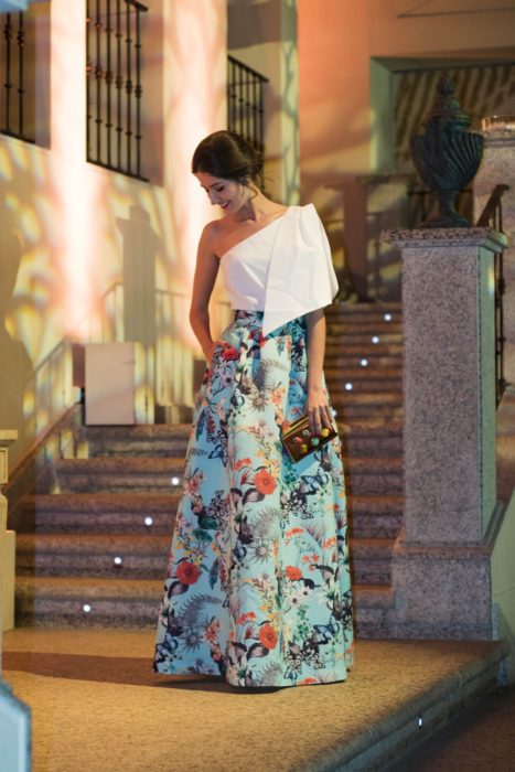 Ideas de vestidos para boda en jardín al aire libre; chica con vestido de fiesta, falda azul floreada, top blanco de un solo hombro y bolsa de mano