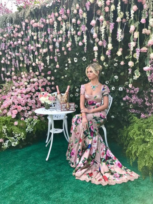 Ideas de vestidos para boda en jardín al aire libre; chica rubia con cabello agarrado en chongo, con vestido rosa floreado, largo hasta el suelo, sentada en un jardín con rosas blancas y rosadas