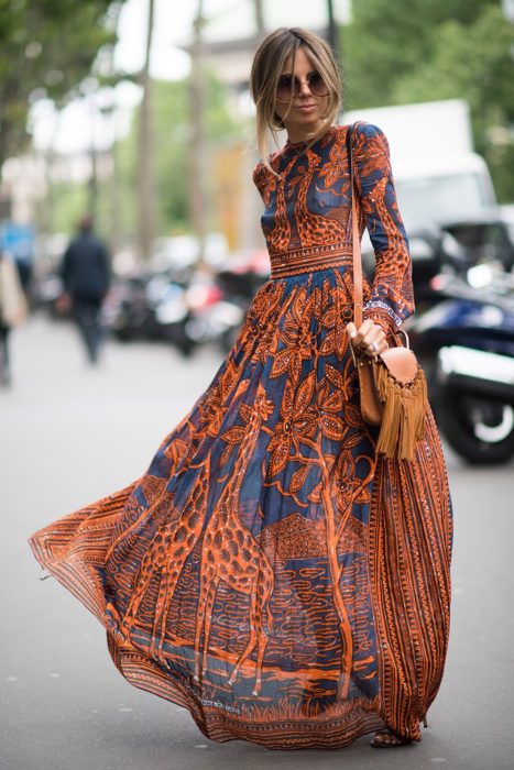 Ideas de vestidos para boda en jardín al aire libre; mujer rubia con peinado despeinado, lentes de sol grandes y redondos, con vestido azul y anarangado, con mangas y largo hasta el suelo, con estampado de árboles y jirafas