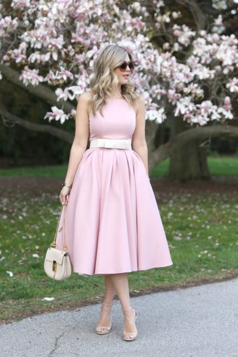 Ideas de vestidos para boda en jardín al aire libre; chica de cabello rubio y ondulado, con lentes de sol, vestido rosa, cinturó blanco, stilettos abiertos y bolsa color hueso, posando frente a un árbol de flores rosas