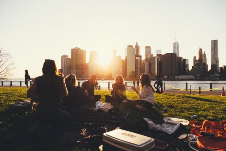 Grupo de jóvenes sentados en en el pasto frente a un lago, mientras estudian y charlan
