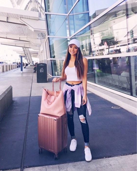 Chica sosteniendo su maleta color rosa fuera del aeropuerto, despidiéndose antes de abordar su vuelo