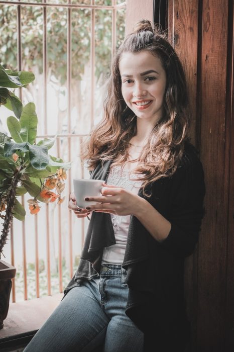 una mujer recargada en una ventana toma una taza de café con las dos manos