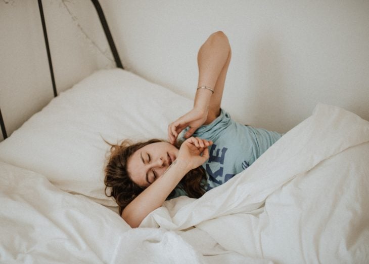 una mujer con blusa azul está estirándose mientras duerme entre sábanas beige