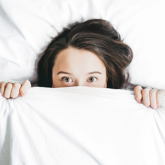 una mujer acostada entre sabanas blancas asoma sus ojos abiertos por encima de la sábana