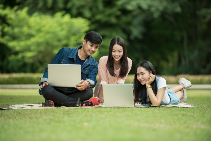 tres jóvenes sentados en el césped ven dos computadoras portátiles