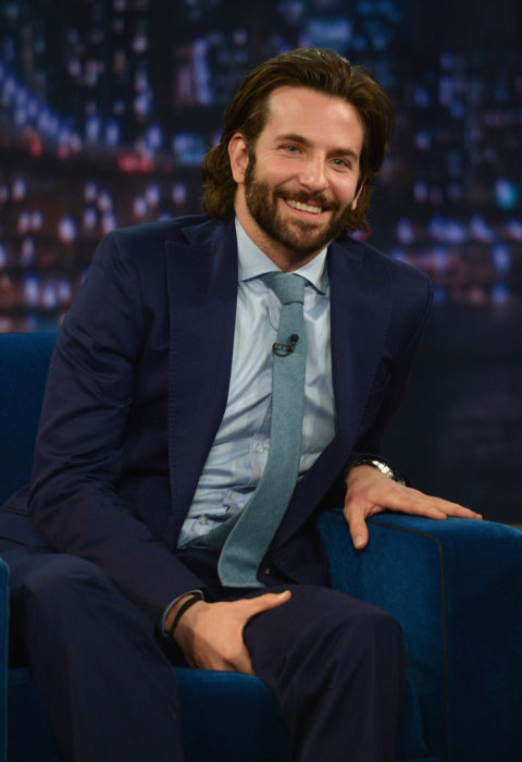 Actor, director y cantante Bradley Cooper con cabello largo y barba, con traje sentado en un sillón