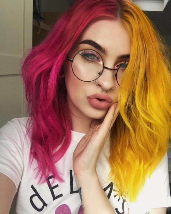 Chica con lentes redondos de aumento, perforaciones en la nariz y tabique, con delineador rosa y cabello teñido de dos colores: rosa magenta y amarillo canario