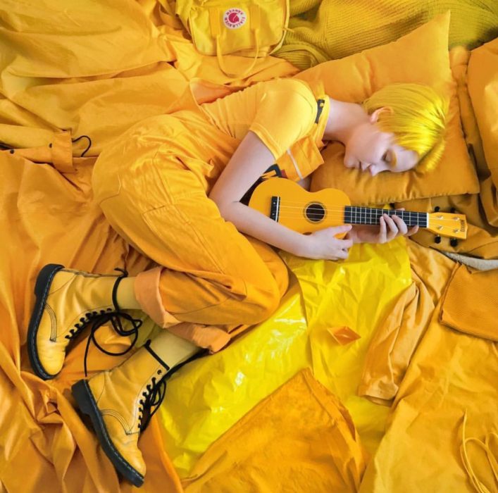 Chica con cabello amarillo y corte pixie, con botas Dr. Martens y ukulele, rodeada de cosas color amarillo