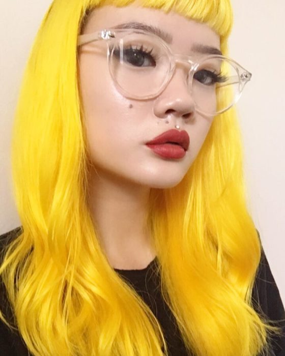 Chica asiática con grandes lentes vintage de aumento, con perforación encima del labio y cabello largo, ondulado y teñido de amarillo brillante