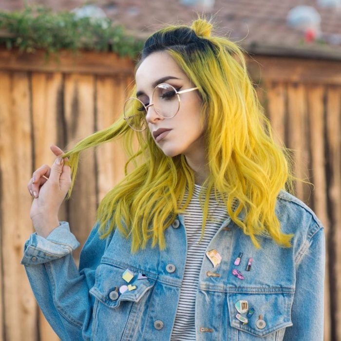 Chica con maquillaje profesional, lentes vintage redondos y grandes, con chaqueta denim y pines, tiene cabello amarillo con las raíces negras