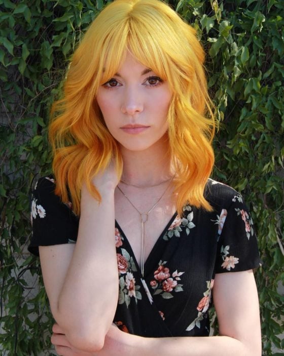 Chica con mirada triste, de cabello amarillo degradado a anaranjado, corte bob con fleco y vestido de flores