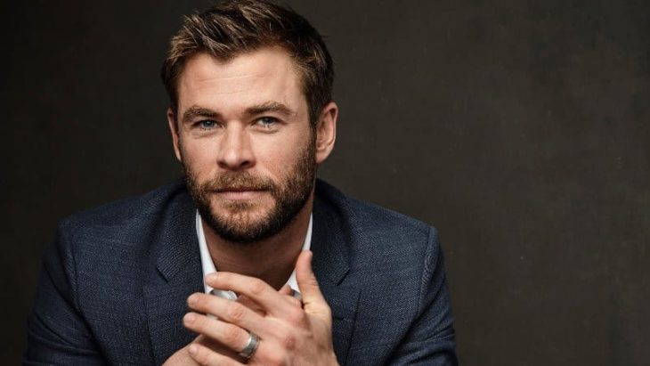 Chris Hemsworth posando para uyna fotografía, usando traje sastre azul marino, mirando al frente y con las palmas de las manos cruzadas