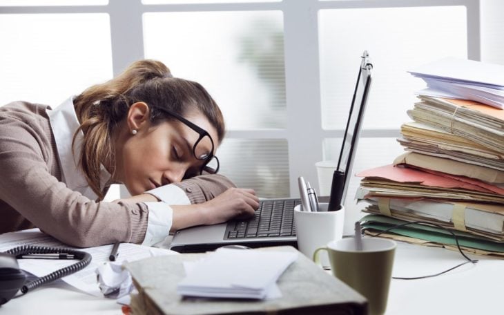 una mujer con lentes está acostada sobre su computadora portátil en una oficina con papeles por todos lados