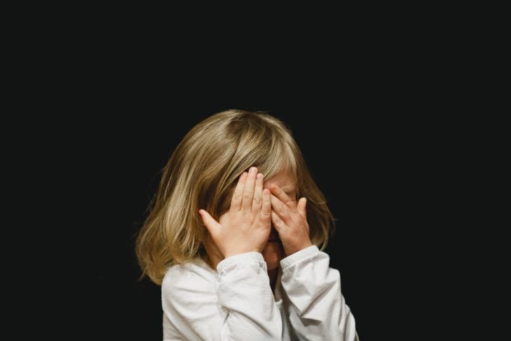 una niña rubia cubre su cara con sus manos sobre un fondo negro