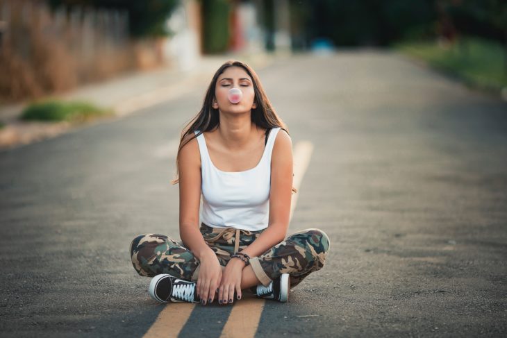 una chica sentada en la calle en posición de flor de loto mastica chicle y hace una pompa