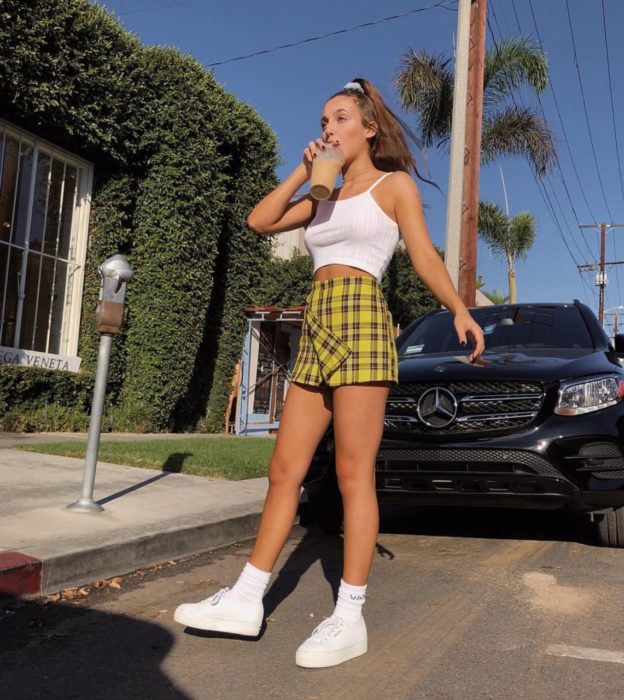 Faldas para el verano; chica en la calle tomando un café helado, con blusa blanca de tirantes, falda de tela escocesa amarilla y tenis blancos