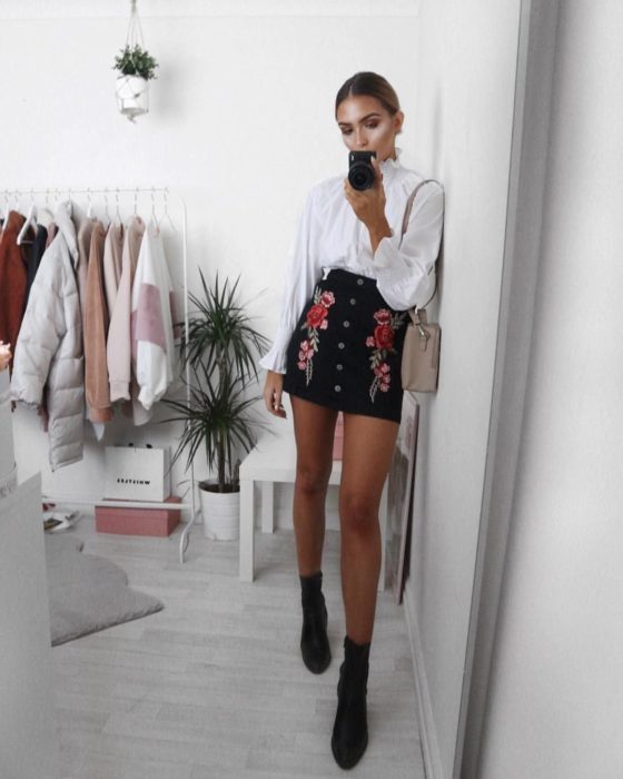 Faldas para el verano; chica tomándose selfie frente al espejo, con blusa blanca, falda negra con rosas bordadas y botines negros