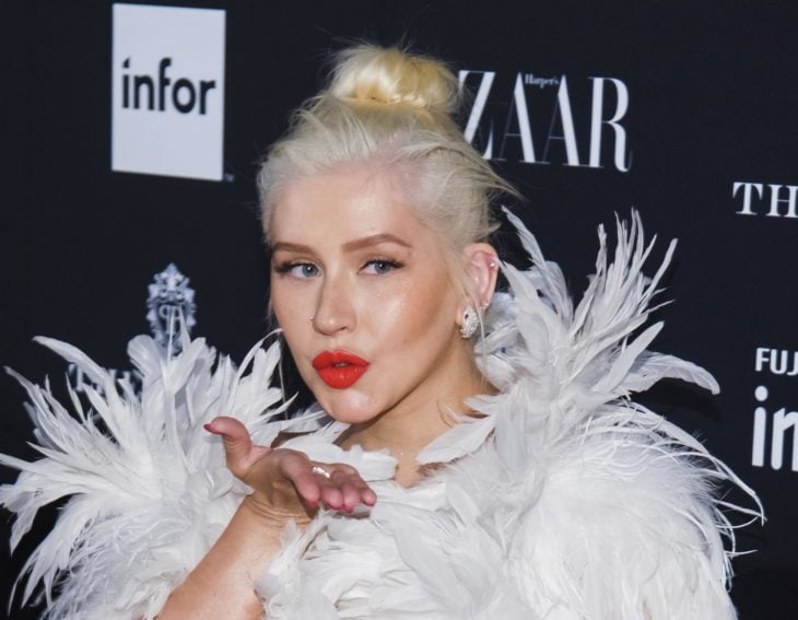 Cantante Christina Aguilera con cabello rubio y peinado con un chongo, usando un vestido de plumas blancas 