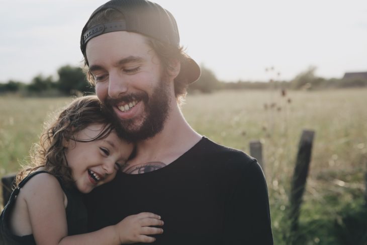 un hombre con cachucha abraza a su niña con el fondo de un campo verde