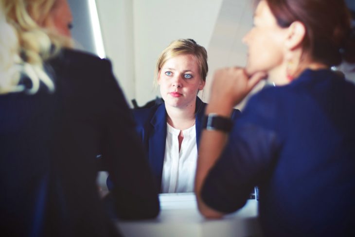 tres mujeres platican en una mesa, dos las vemos de espaldas parcialmente y una en medio al frente