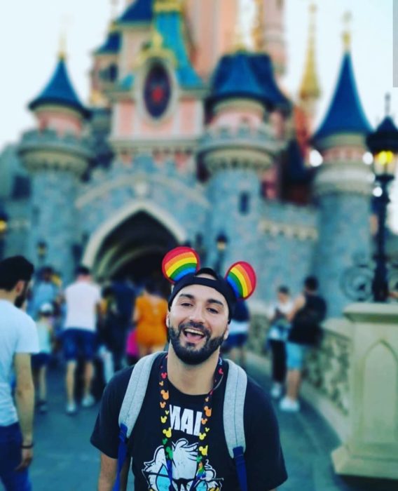 Magical Pride en Disneylandia; chico con barbay cachucha de orejas de Mickey Mouse frente a castillo 