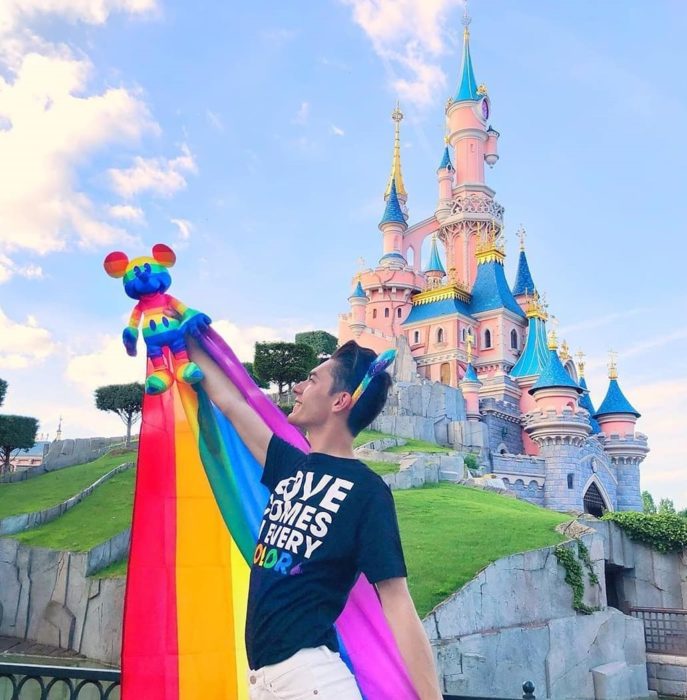 Magical Pride en Disneylandia; chico frente al castillo Disney sosteniendo una bandera de colores y un muñeco de Mickey Mouse