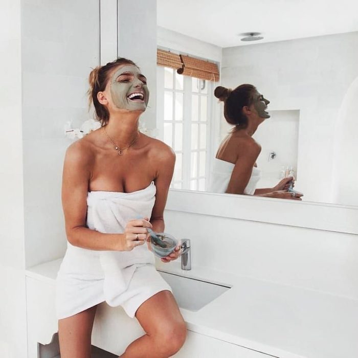 Chica con una toalla enredada, sobre un lavabo, aplicando una mascarilla verde