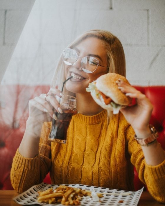 Chica bebiendo soda y sosteniendo una hamburguesa dentro de un restaurante