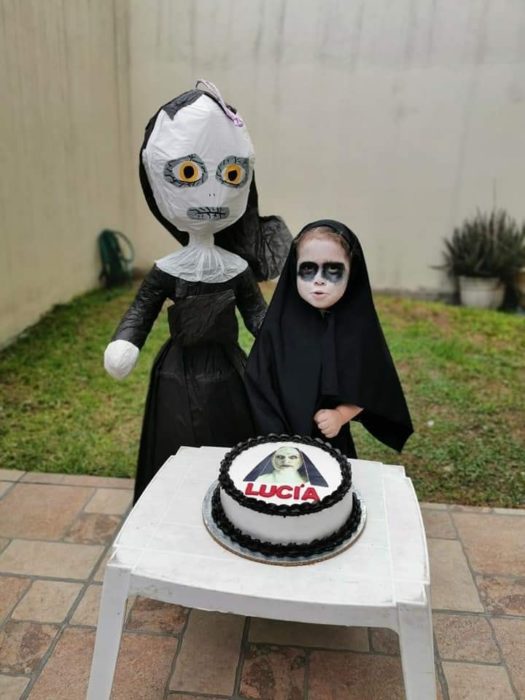 Niña frente a un pastel de cumpleaños, en un jardín, disfrazada de La Monja 