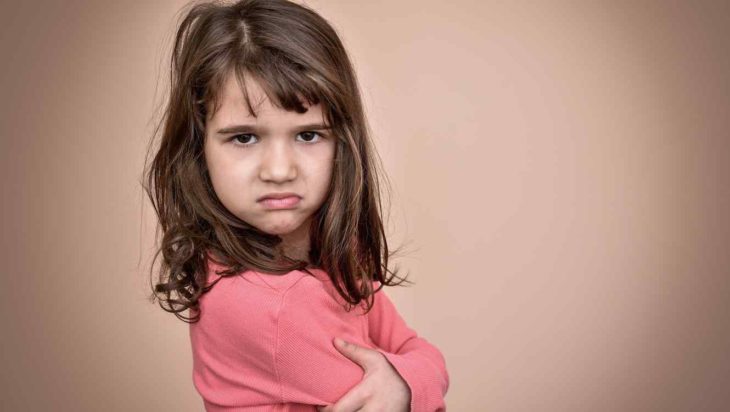 una niña de pelo castaño hasta los hombros y con brazos cruzados mira a la cámara con aspecto enojado