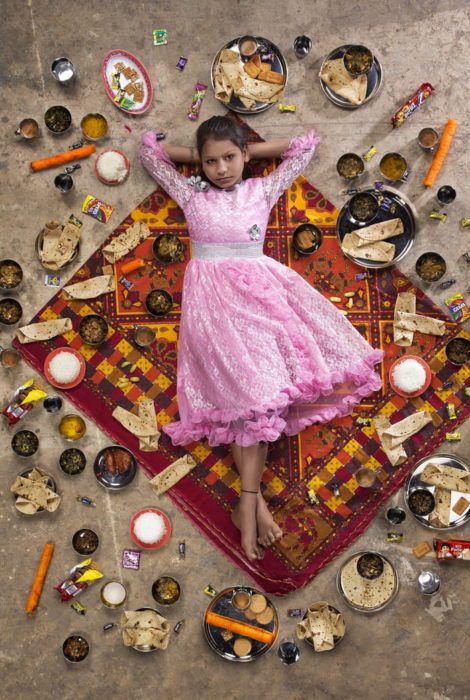 Niña con vestido rosado, recostada sobre una alfombra roja, rodeada de comida, proyecto fotográfico de Gregg Segal