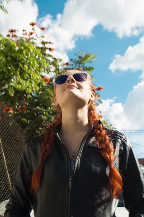 una mujer pelirroja con trenzas y lentes sonríe en una toma contrapicada