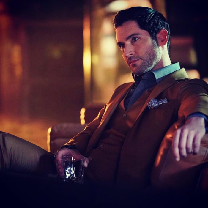 Actor de serie de Netflix, Lucifer, Tom Ellis; hombre de traje café sentado en un sillón sosteniendo un vaso 