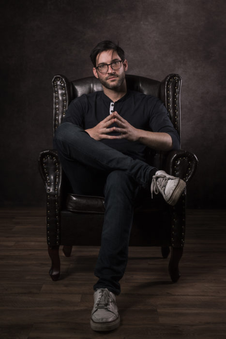 Actor de serie de Netflix, Lucifer, Tom Ellis; hombre de barba y lentes sentado en sillón de cuero con las piernas cruzadas 