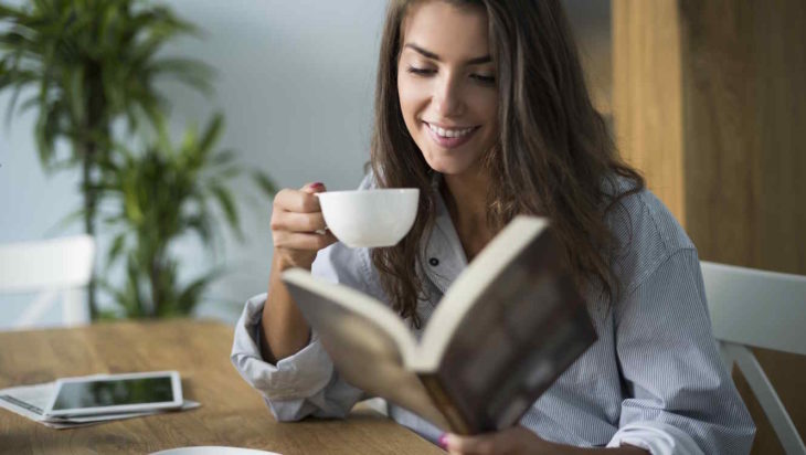 una mujer lee un libro y toma una taza de café