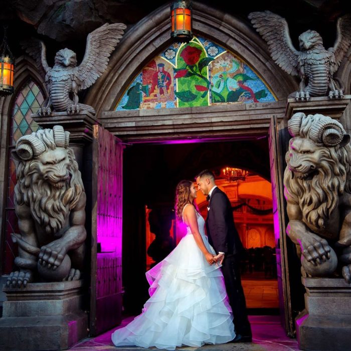 Precios de boda en Disneylandia; pareja de recién casados tomados de la mano frente al castillo de La Bella y la Bestia, con gárgolas y estatuas de animales mitológicos