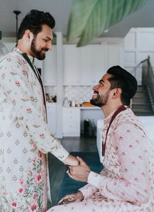 Pareja gay de la india celebrando su boda al estilo hindú. Chico de rodillas sosteniendo la mano de su esposo 